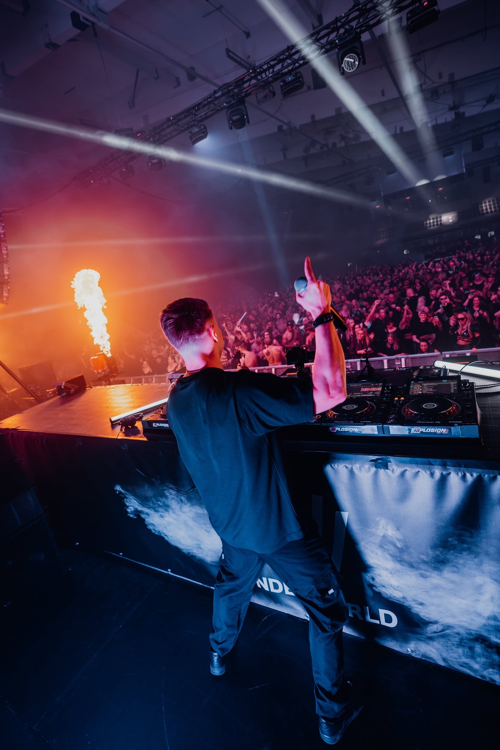 TISCORE raising a microphone behind the decks, a packed arena crowd in front of him and a flame jet beside the stage.