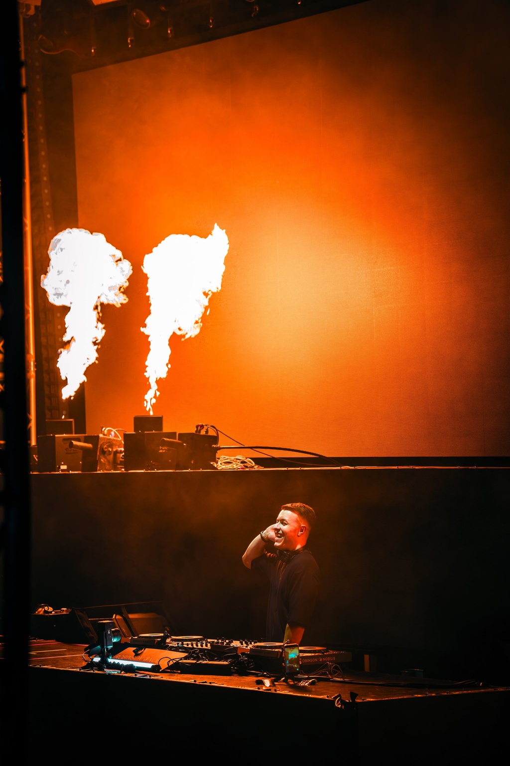 TISCORE behind the decks with two flame jets and an orange lit stage wall behind him.